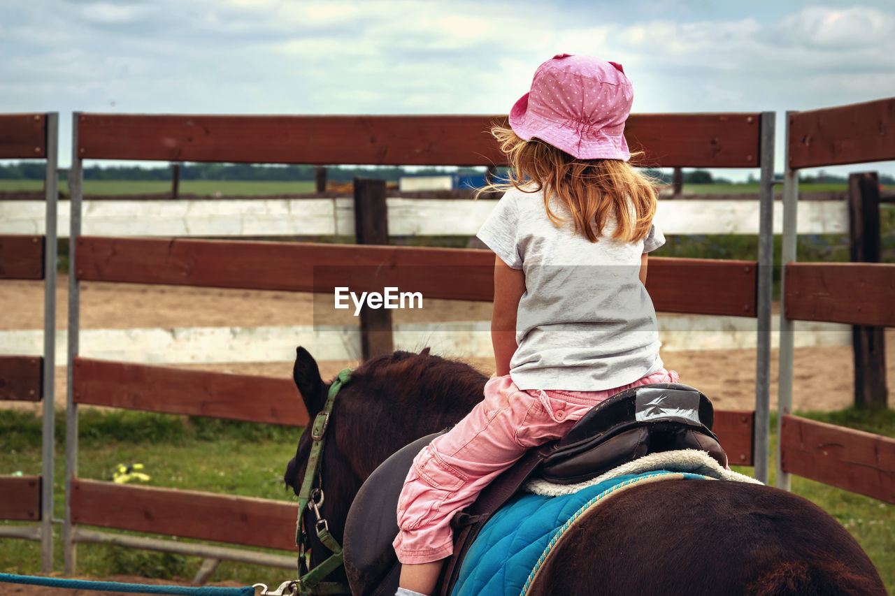 REAR VIEW OF WOMAN SITTING ON HORSE IN RANCH