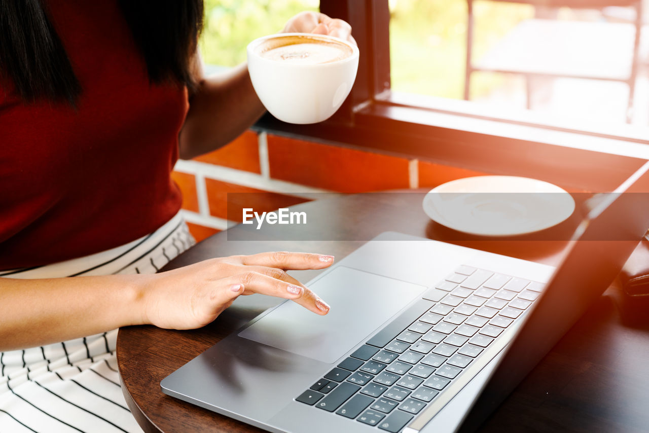 Asia woman hold cup of coffee while typing on laptop keyboard. w