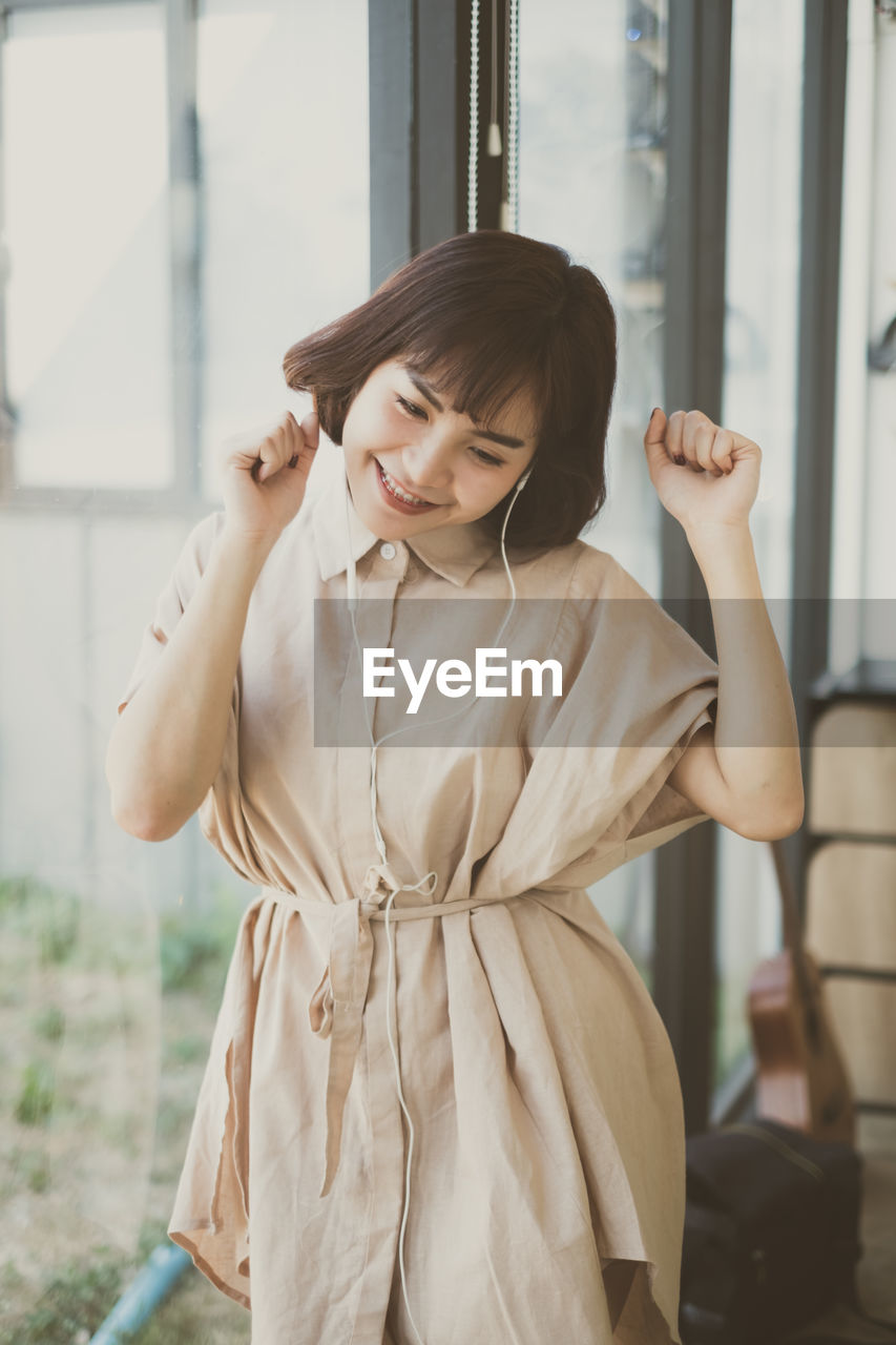 Smiling young woman listening music while standing by window at home