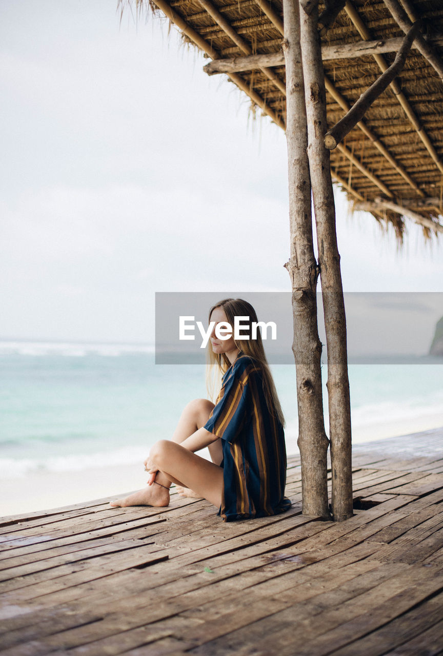 Young woman sitting on shore at beach against sky