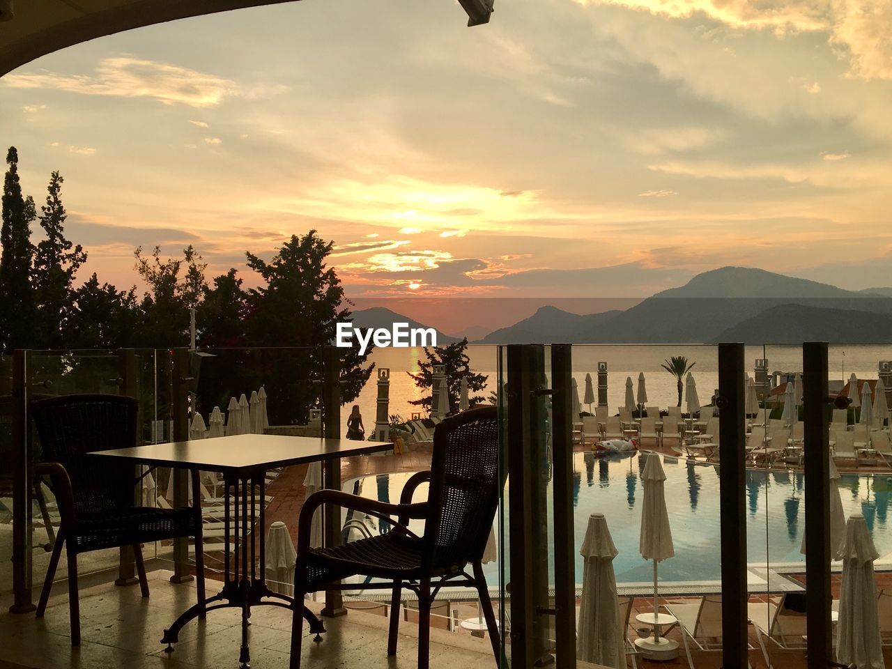 CHAIRS AND TABLE BY SWIMMING POOL AGAINST SKY AT SUNSET