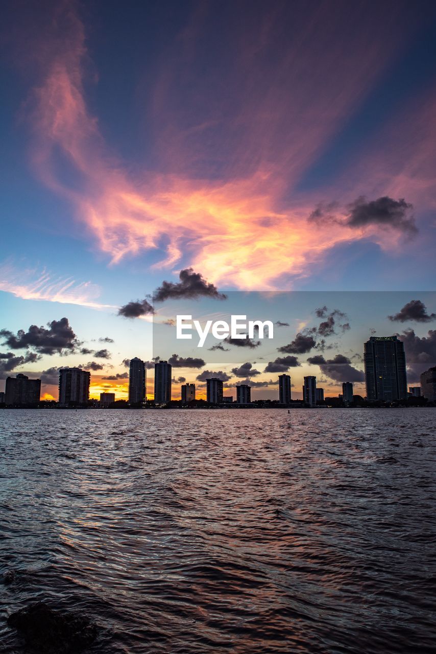 Sea by cityscape against sky during sunset