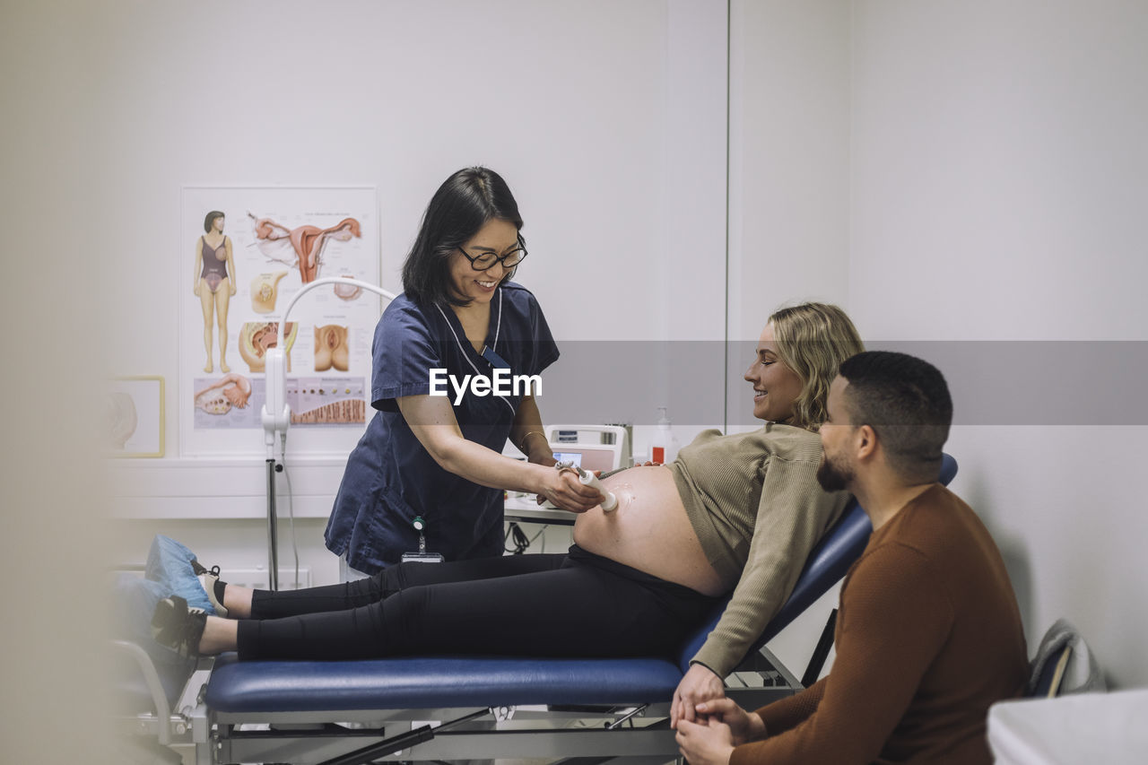 Smiling gynecologist doing ultrasound of pregnant woman sitting by man at hospital