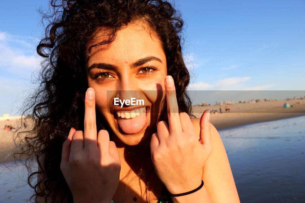 Close-up portrait of cheerful young woman showing obscene gesture at beach