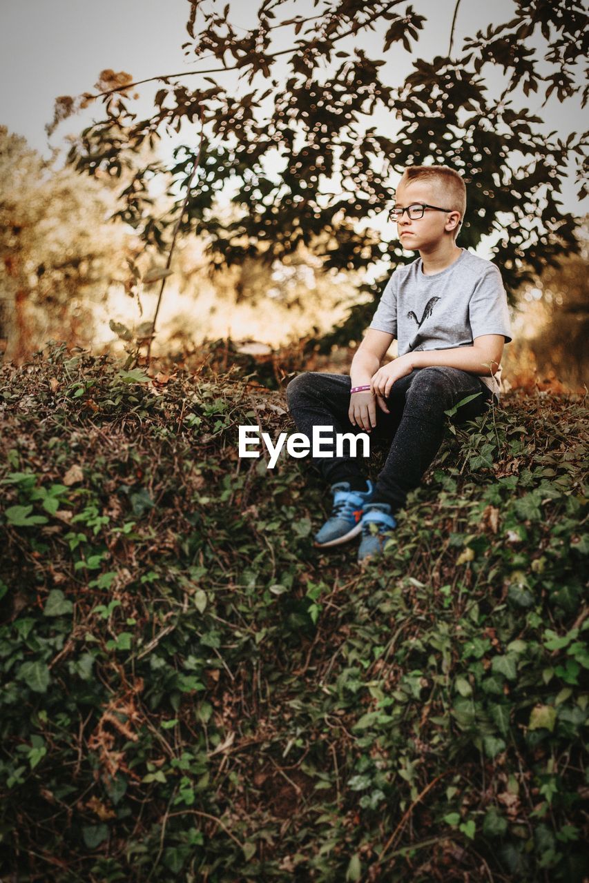 FULL LENGTH OF YOUNG MAN SITTING ON PLANT AT FIELD