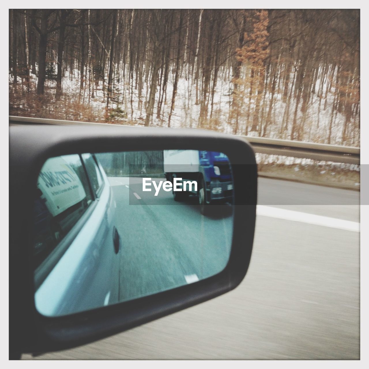 Reflection of car in side-view mirror against road and trees during winter