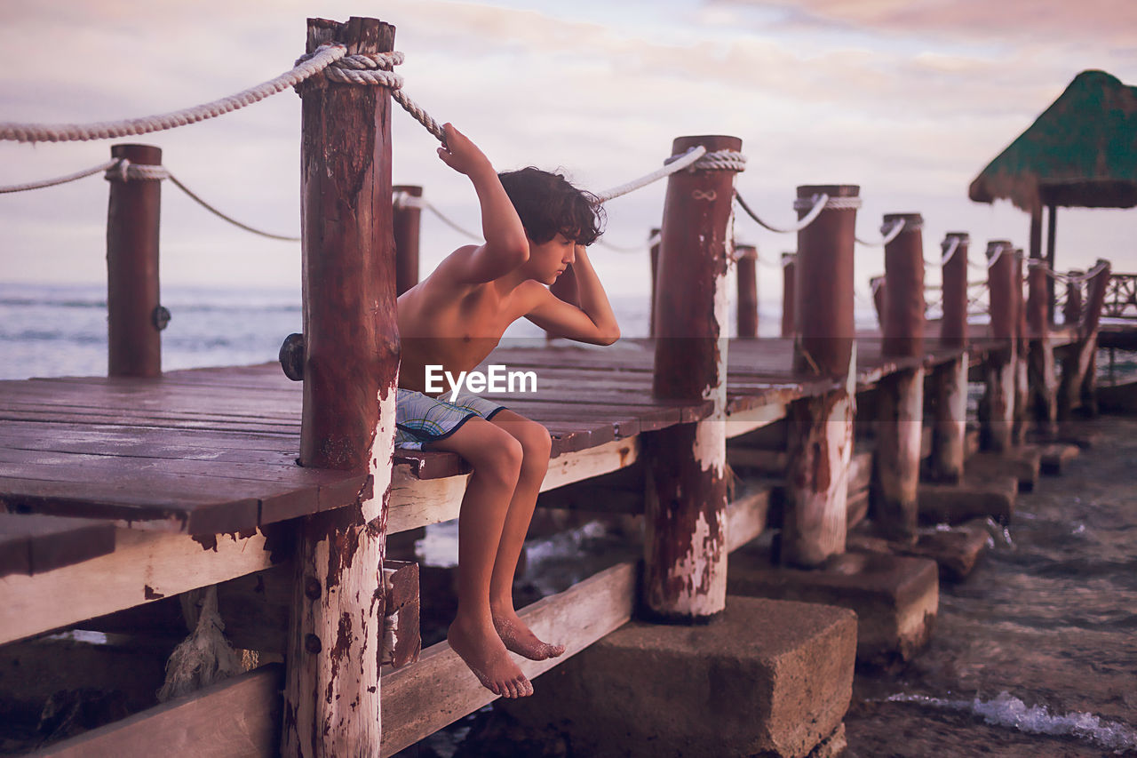 Pre teen boy sitting on a wooden pier at a tropical beach.