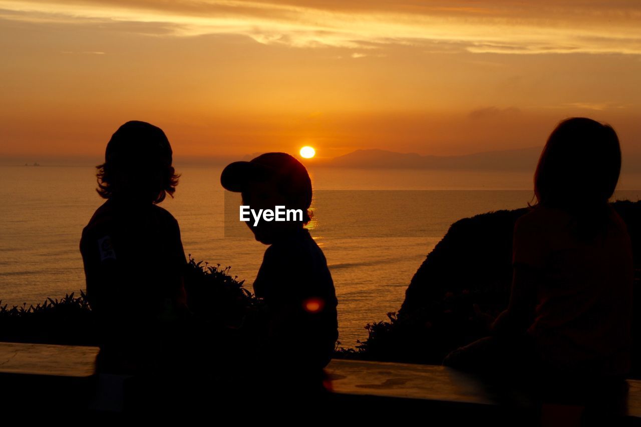 SILHOUETTE PEOPLE SITTING ON SHORE AGAINST ORANGE SKY