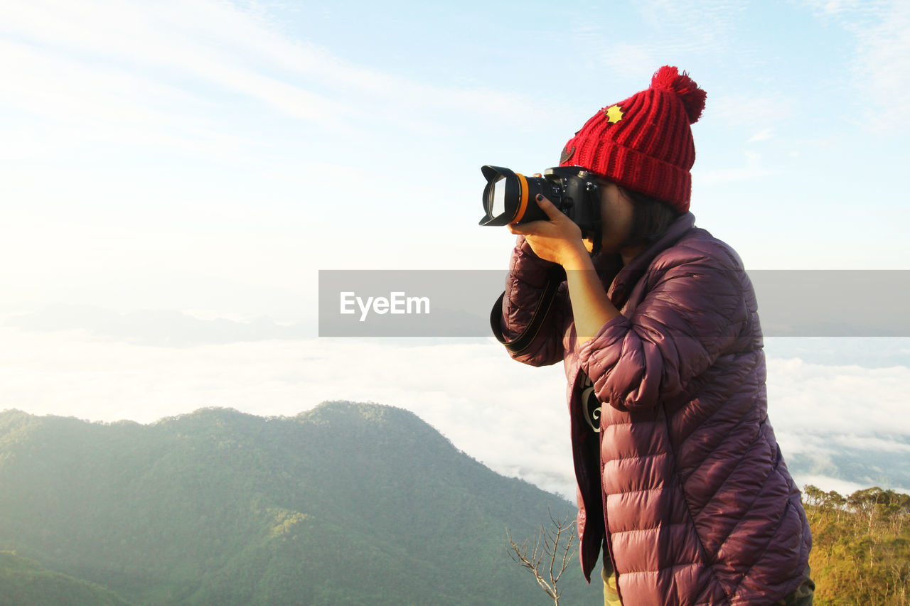 Side view of woman wearing jacket photographing with digital camera against mountains