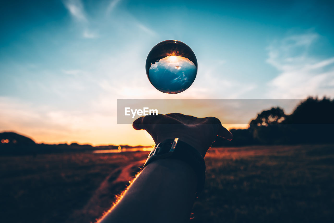 Cropped man catching crystal ball against sky during sunset