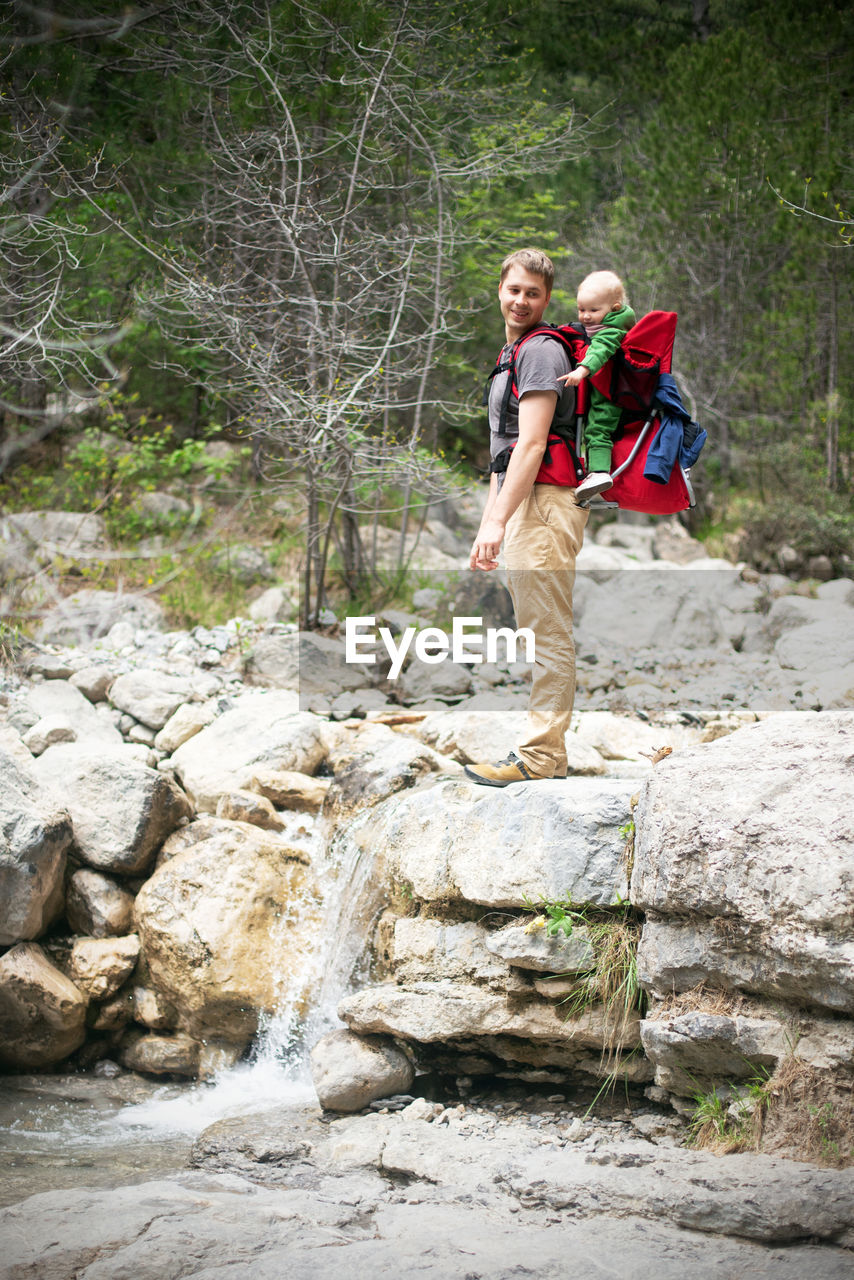 Young man hiking with his baby sitting in the backpack. family is watching small waterfall in forest