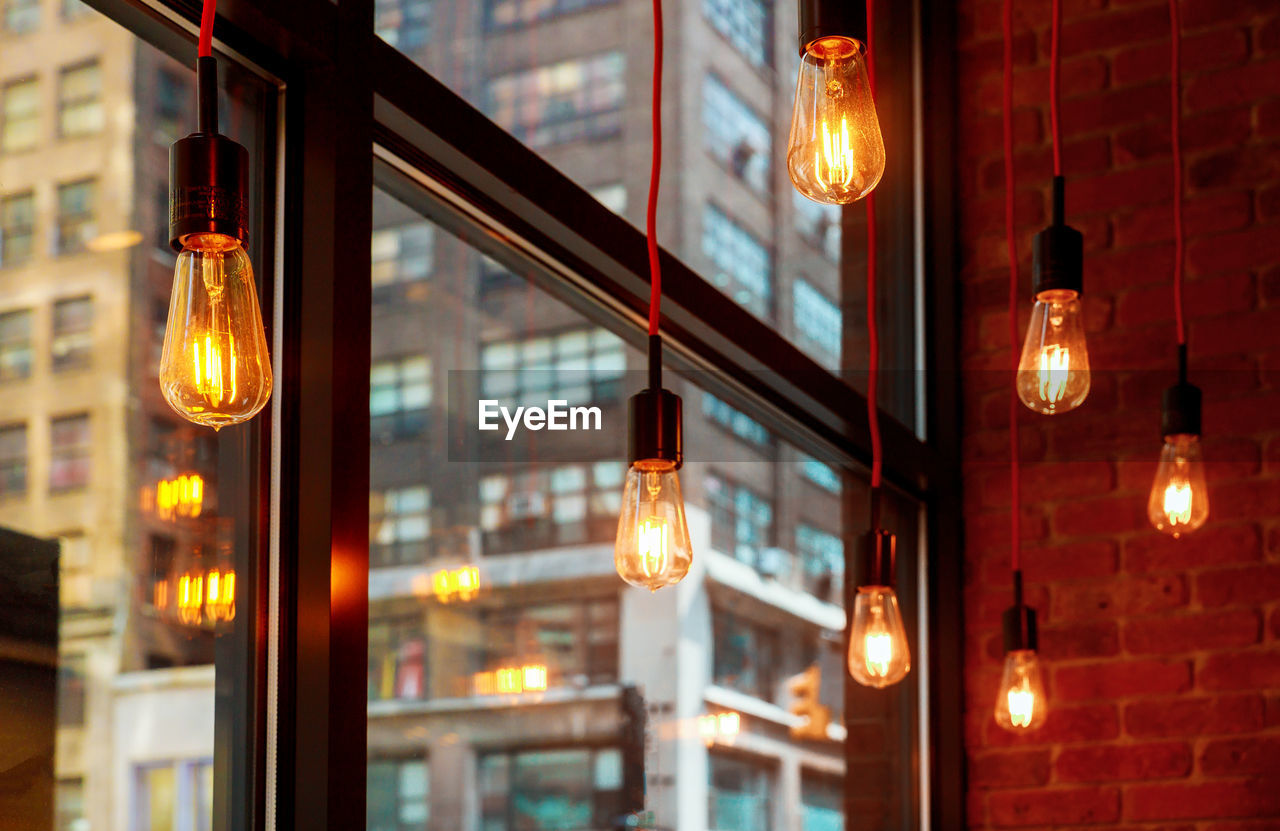 Low angle view of illuminated light bulbs hanging at ceiling by window