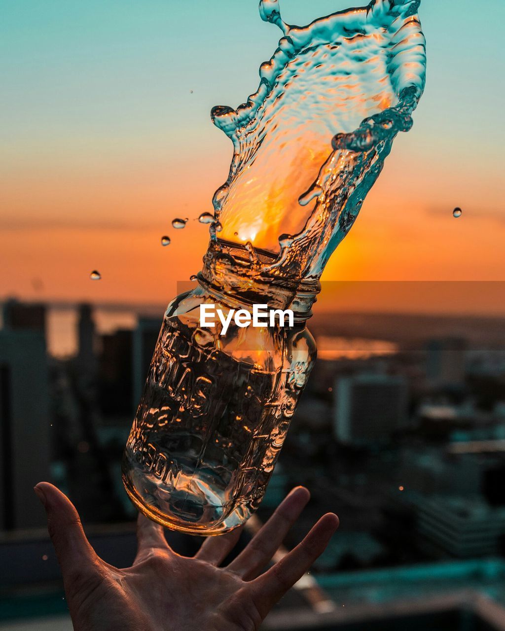 Cropped image of hand catching bottle with splashed water against sky during sunset
