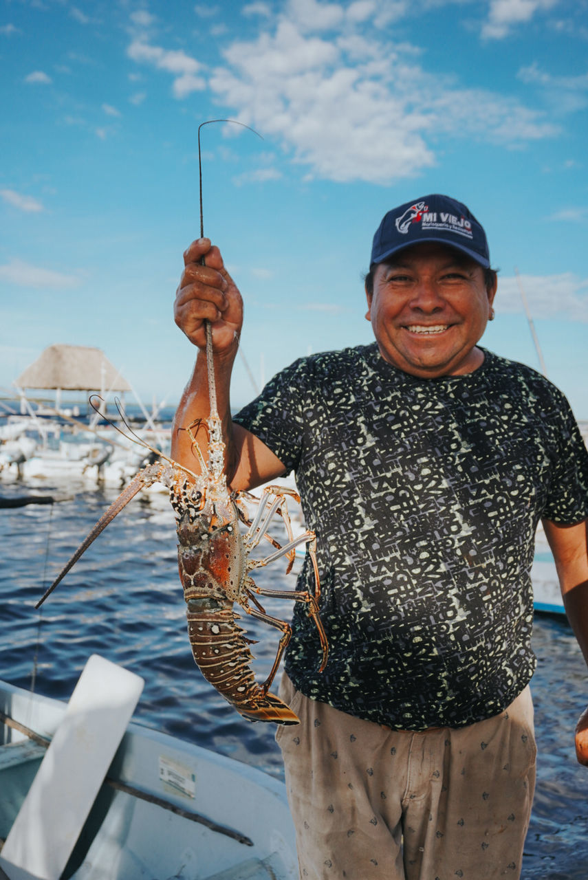 water, fishing, smiling, nautical vessel, looking at camera, adult, one person, sea, portrait, happiness, men, transportation, nature, sky, emotion, front view, standing, senior adult, fish, mode of transportation, mature adult, cheerful, big-game fishing, day, ship, leisure activity, travel, seniors, holiday, vacation, holding, trip, three quarter length, outdoors, sailing, cloud, person, waist up, sailor, sailboat, smile, lifestyles, hat, teeth, casual clothing, travel destinations