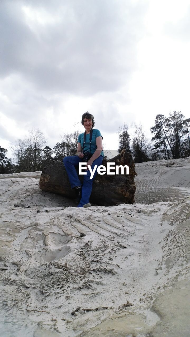 Full length of woman sitting on wood at sandy beach against sky