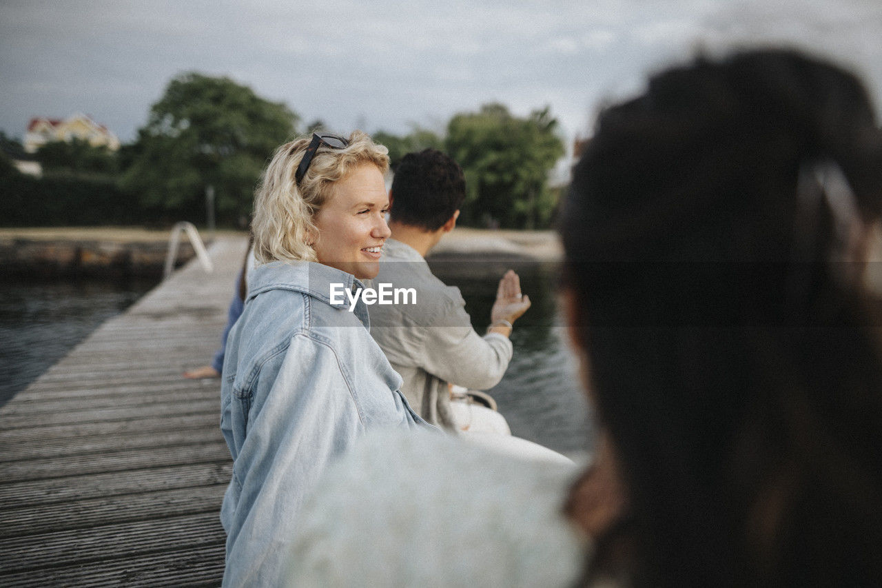 Smiling woman looking away while sitting with friend on pier near lake