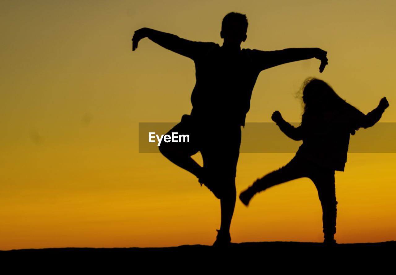 Silhouette siblings dancing on field against sky during sunset