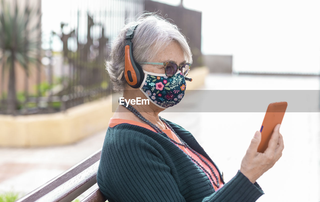 Close-up of senior woman wearing mask listening music while sitting outdoors