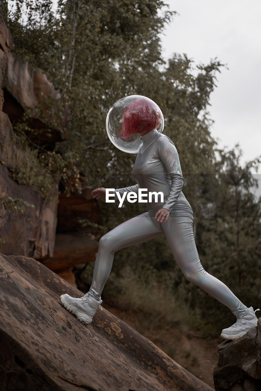 Low angle of positive young female with dyed red hair wearing silver space suit and glass helmet and looking away while walking on rocky formation