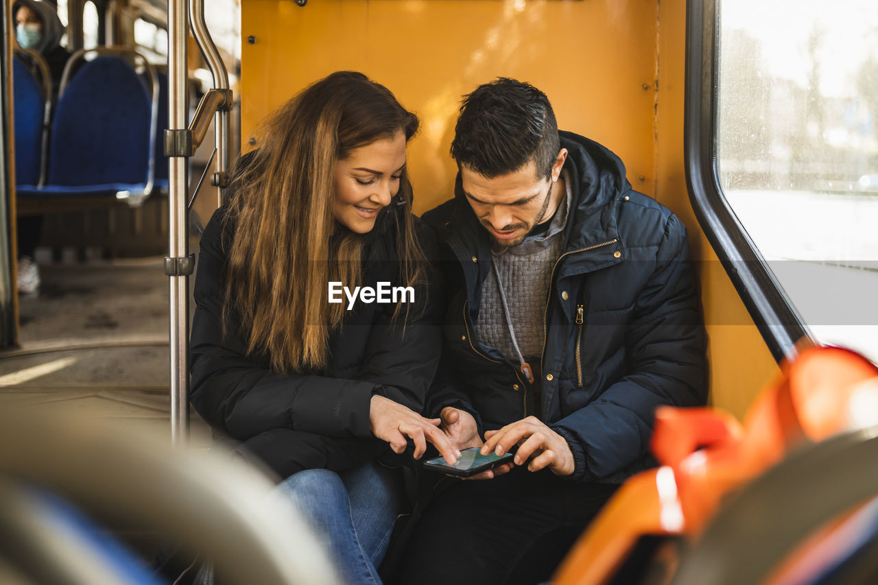 Heterosexual couple using smart phone while sitting in tram during pandemic