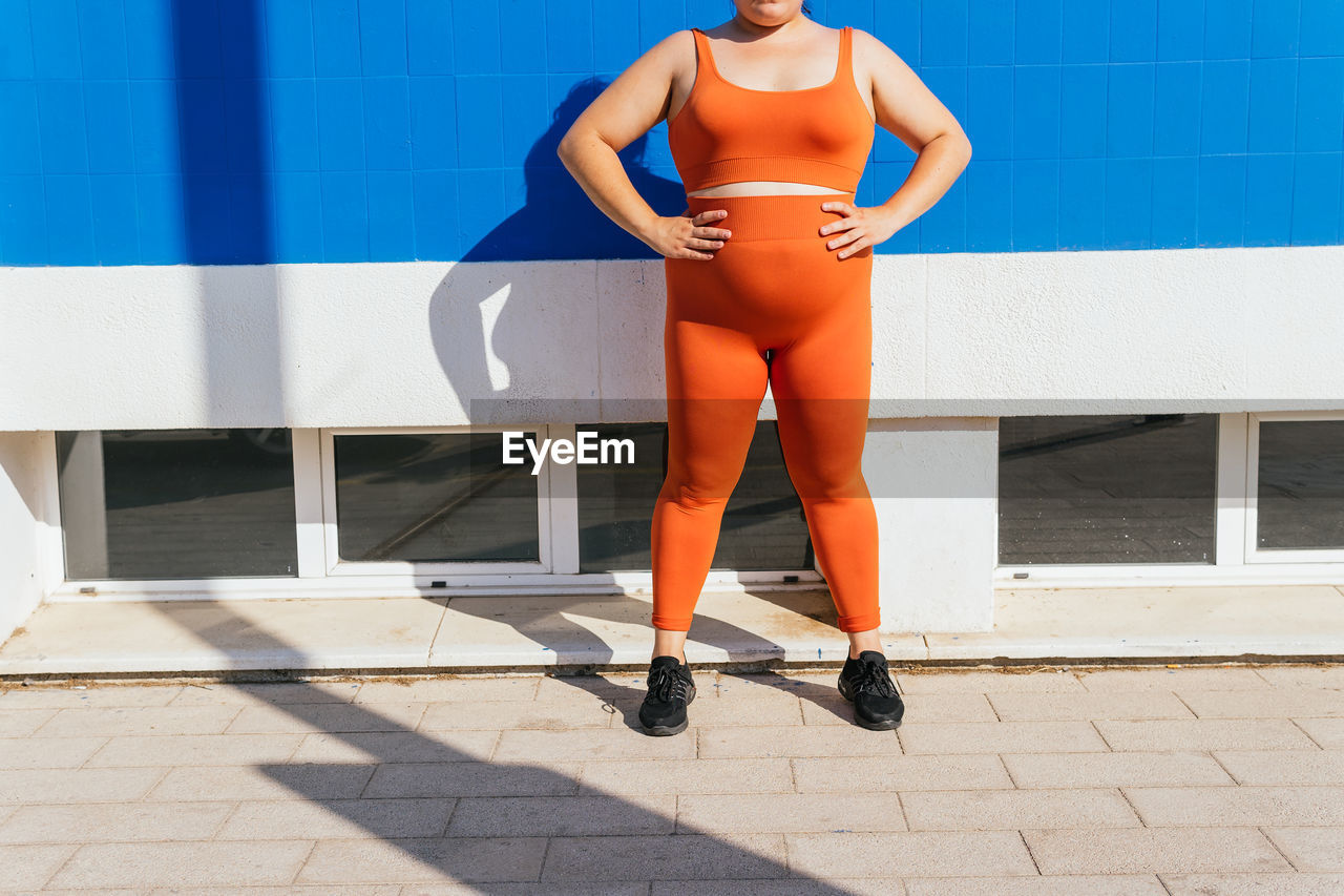 Anonymous overweight ethnic female athlete in active wear against blue tiled wall on sunny day