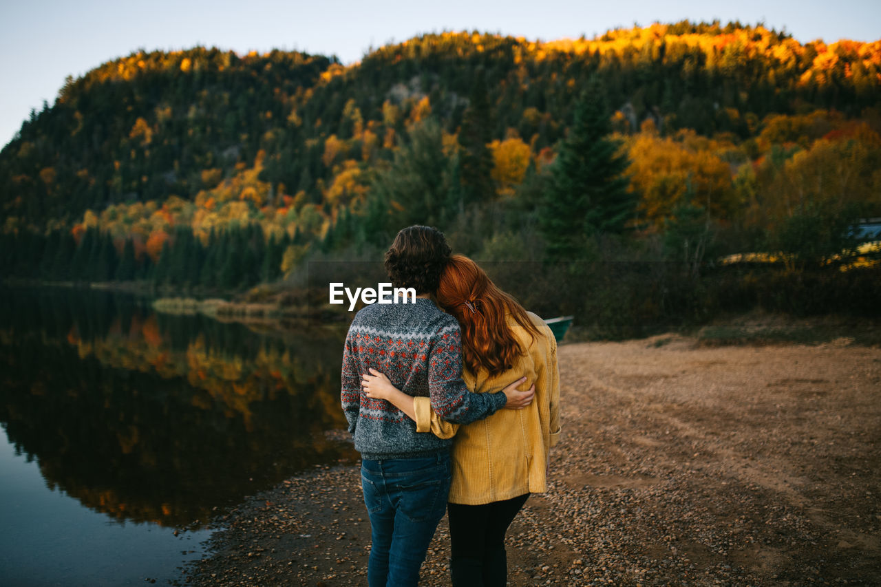 Back view of romantic unrecognizable traveling couple in stylish clothes embracing and admiring scenic autumn landscape of calm lake surrounded by lush forest during trip in canada