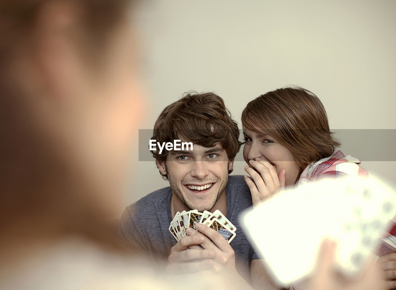 Woman whispering in young man's ear while playing cards with friend at home