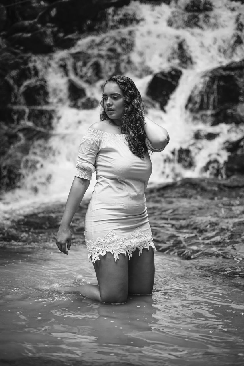 FULL LENGTH PORTRAIT OF YOUNG WOMAN STANDING AGAINST WATER