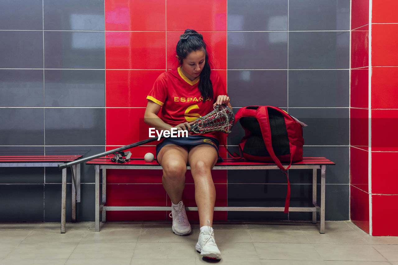 Young athlete adjusting string of lacrosse stick head in locker room