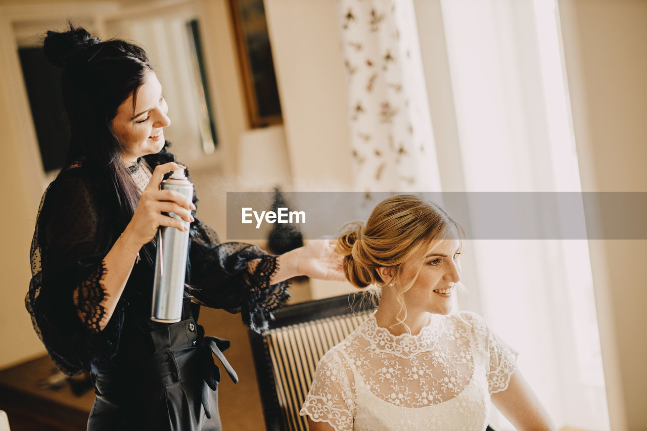 Hairdresser holding hair spray bottle while beautiful bride sitting in wedding dress