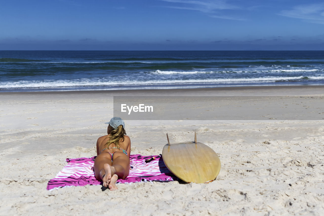 Rear view of woman in bikini lying by surfboard on blanket at beach against blue sky during sunny day