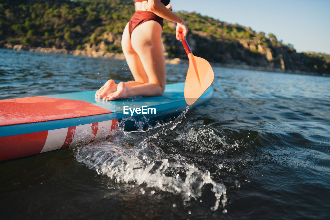 Crop female in swimwear floating on paddleboard in rippling lake against rock and shore with forest on summer day