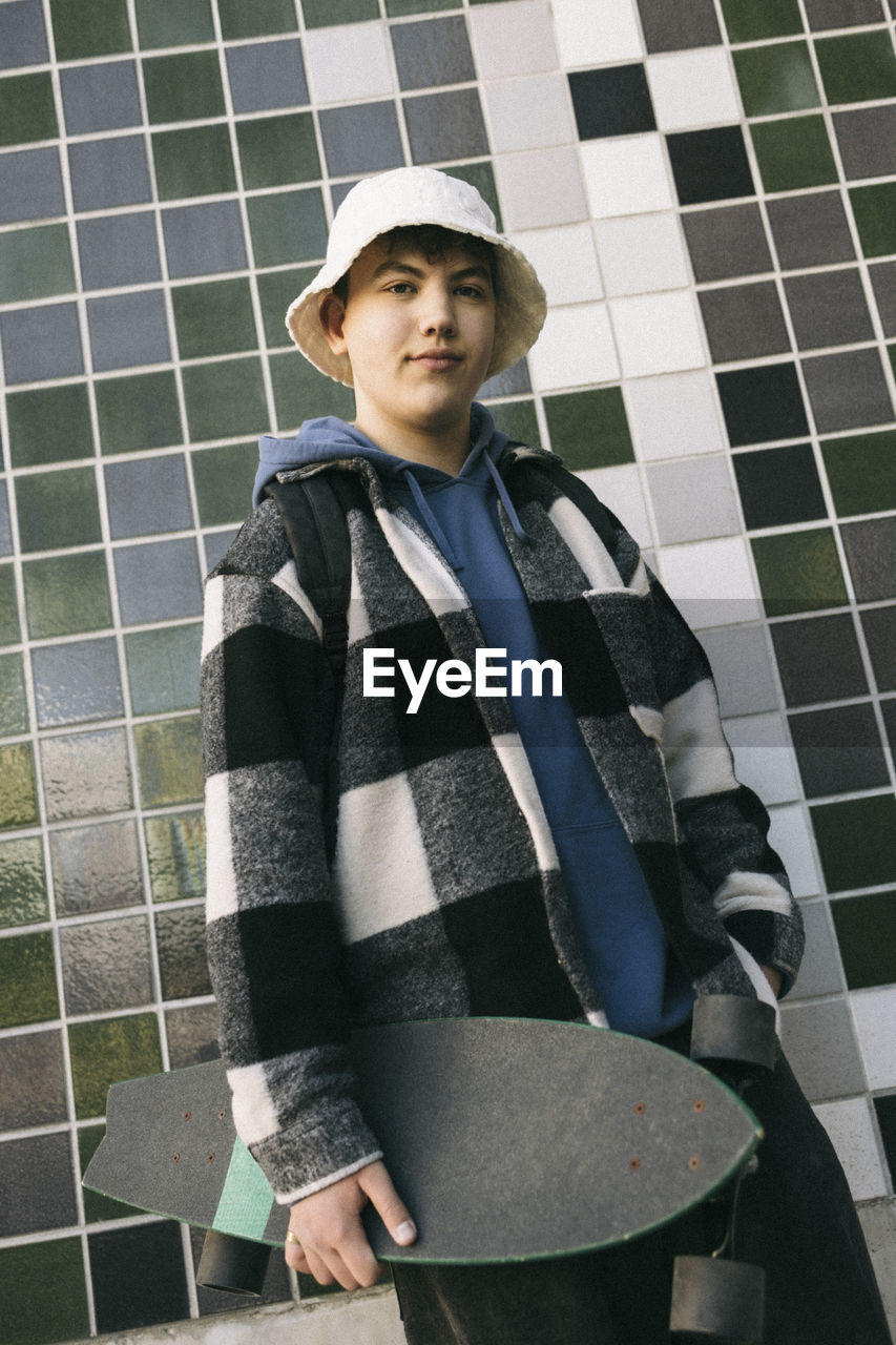 Portrait of teenage boy with skateboard standing against tiled wall