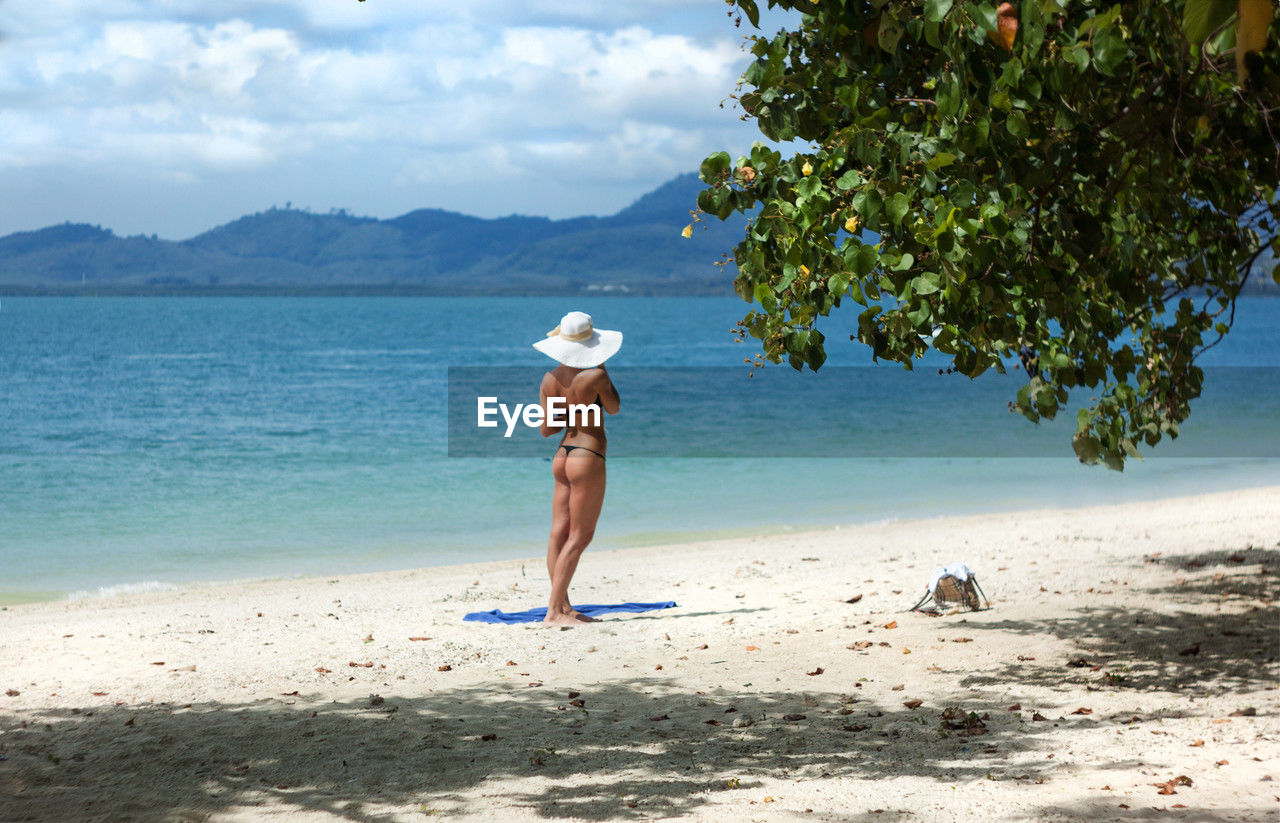 Rear view of sexy slim woman standing at tropical beach against sky and sea