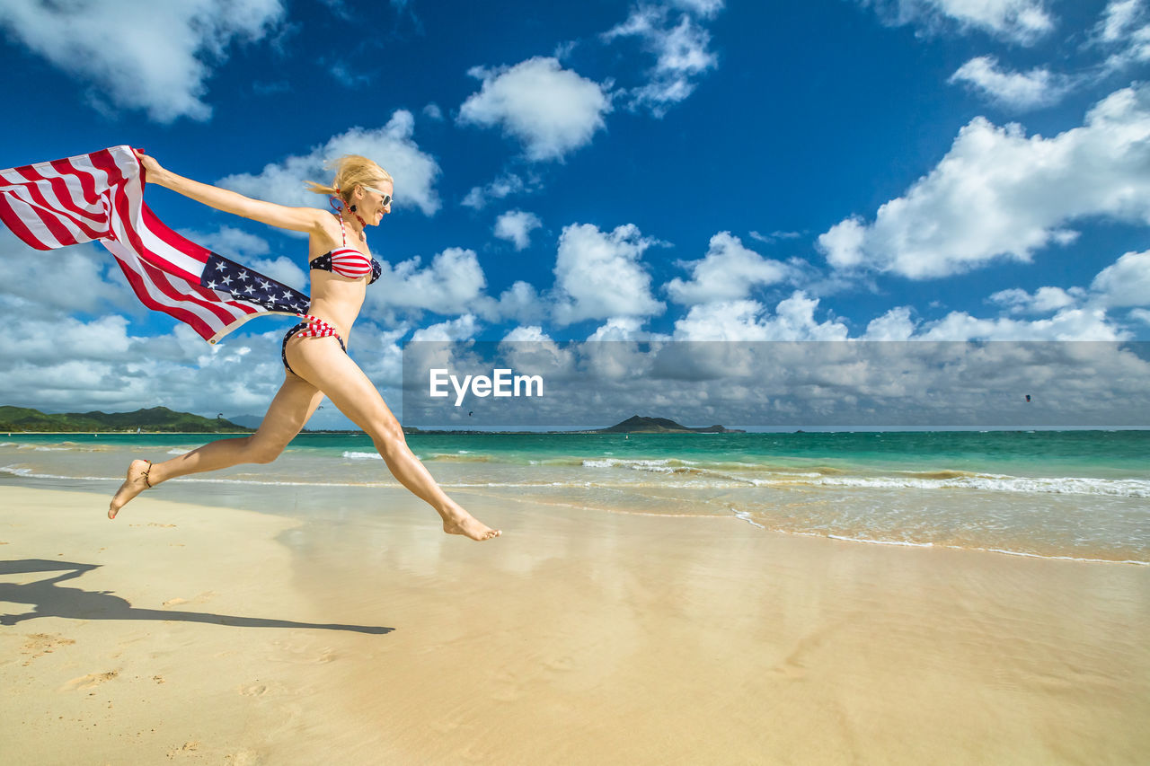 Side view of woman holding american flag while jumping at beach against cloudy sky