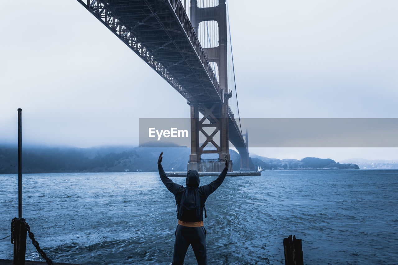 Rear view of man standing under bridge by bay against sky