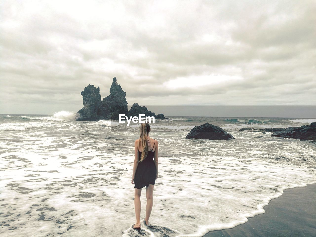 Rear view of young woman standing on shore against cloudy sky