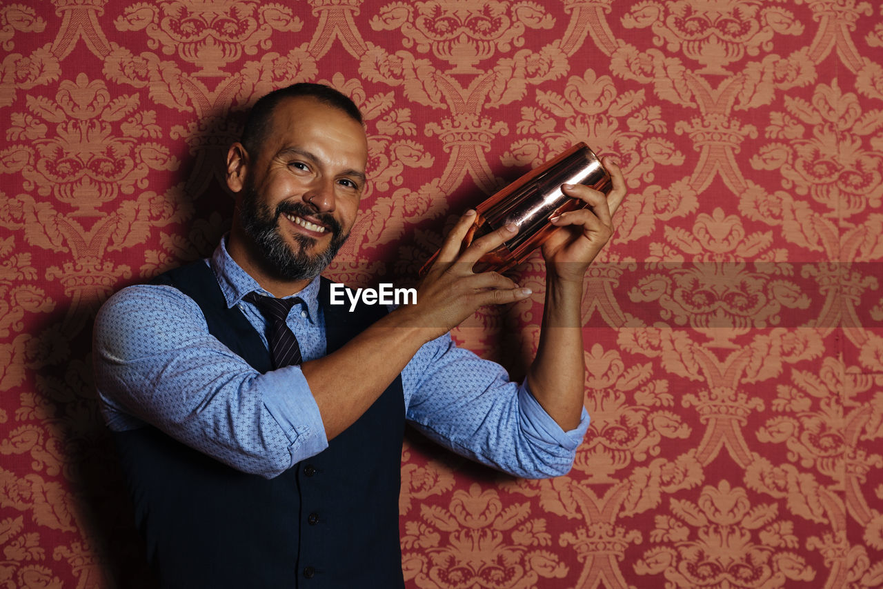 PORTRAIT OF SMILING MAN HOLDING CAMERA WHILE STANDING ON WALL