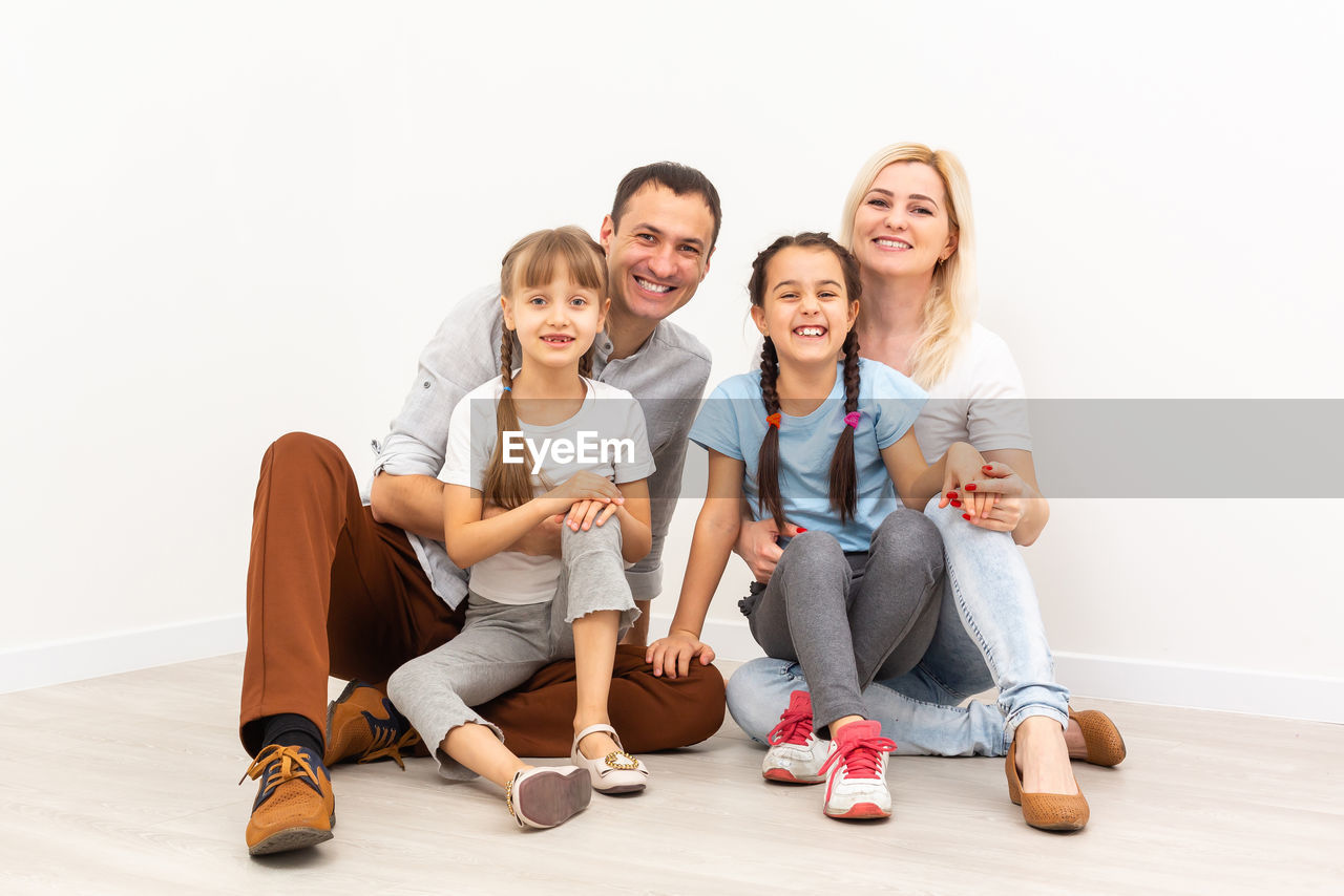 PORTRAIT OF HAPPY FRIENDS SITTING ON FLOOR AGAINST WALL