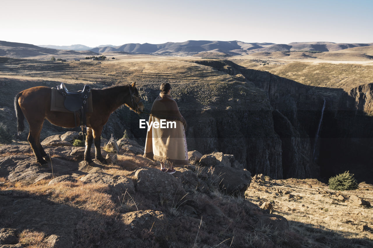 Rear view of woman standing by horse on mountain against clear sky