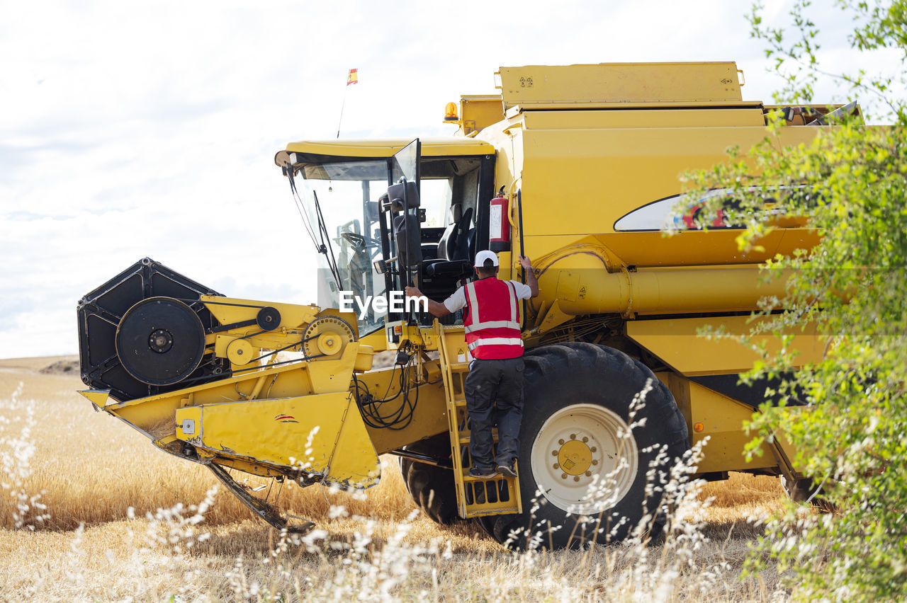 Full body back view of unrecognizable male worker in uniform getting into industrial combine harvester while working in agricultural field