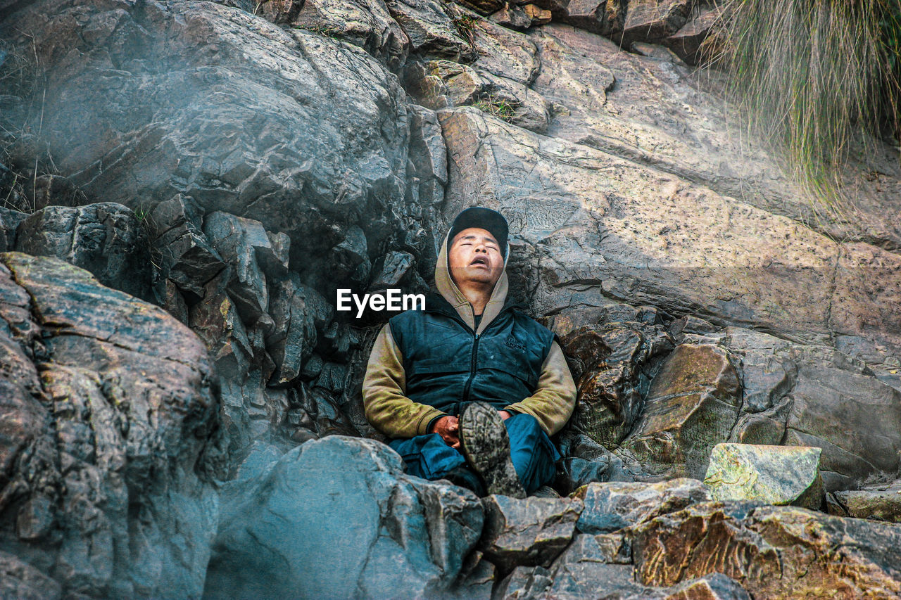 YOUNG MAN SITTING ON ROCK AGAINST WATERFALL