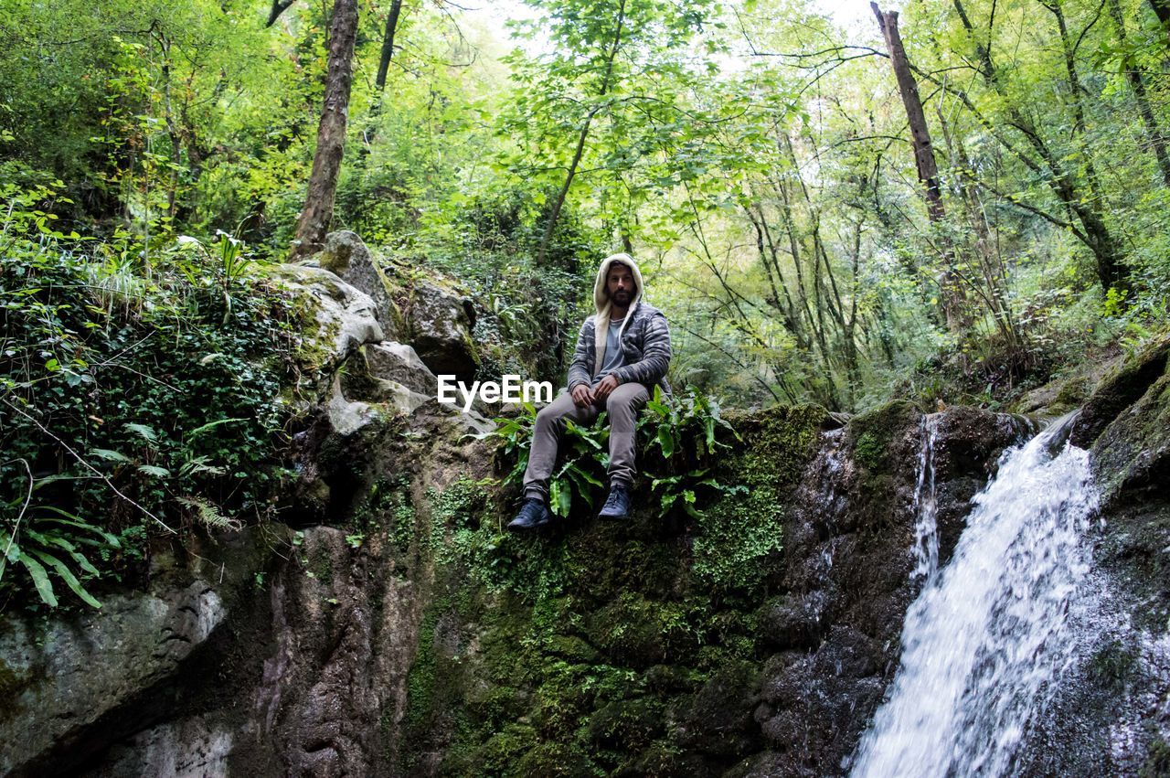 Full length of man sitting by waterfall at forest