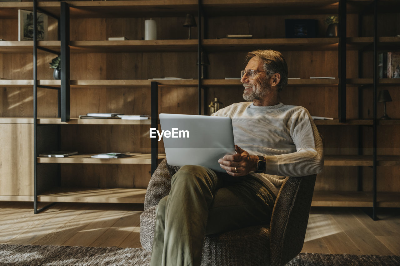 Smiling mature man with laptop sitting on armchair