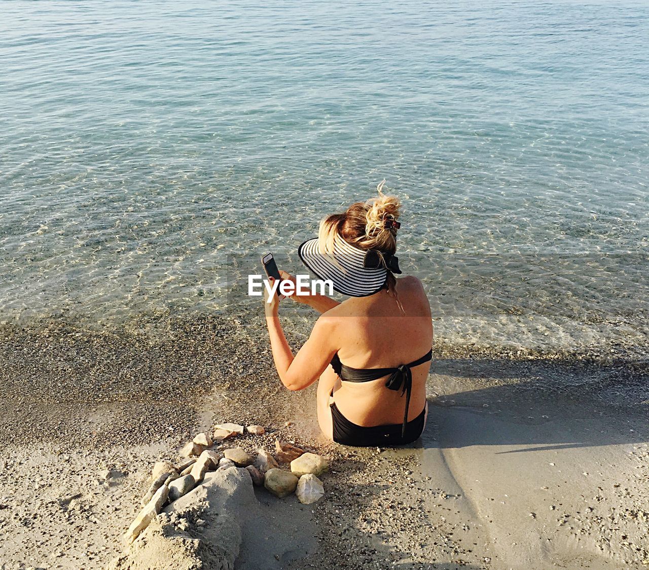 Rear view of woman sitting while using mobile phone on shore at beach