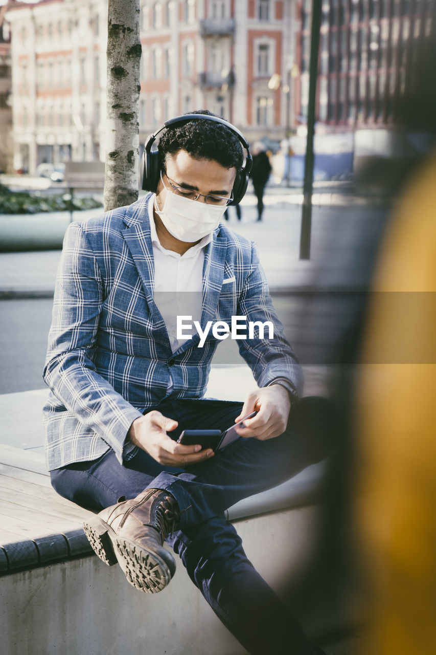 Businessman with smartphone sitting outside with face mask