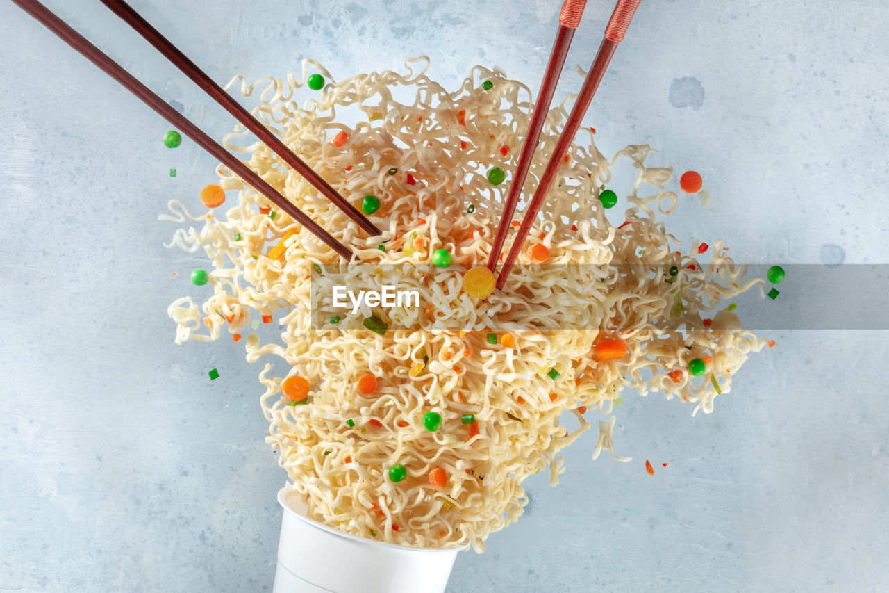 Ramen cup with noodles flying out of it and two pairs of chopsticks, instant noodles