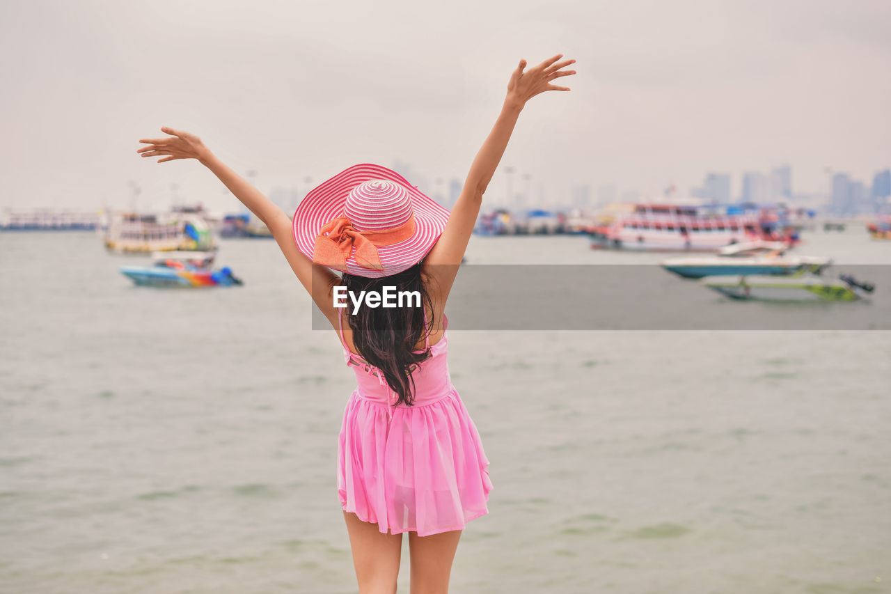 Rear view of woman wearing hat with arms outstretched while standing at beach