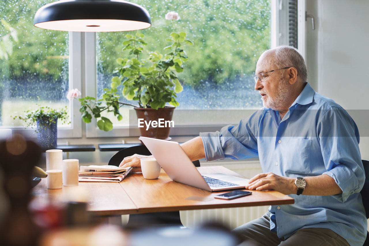 Senior man with laptop reaching for newspaper at table