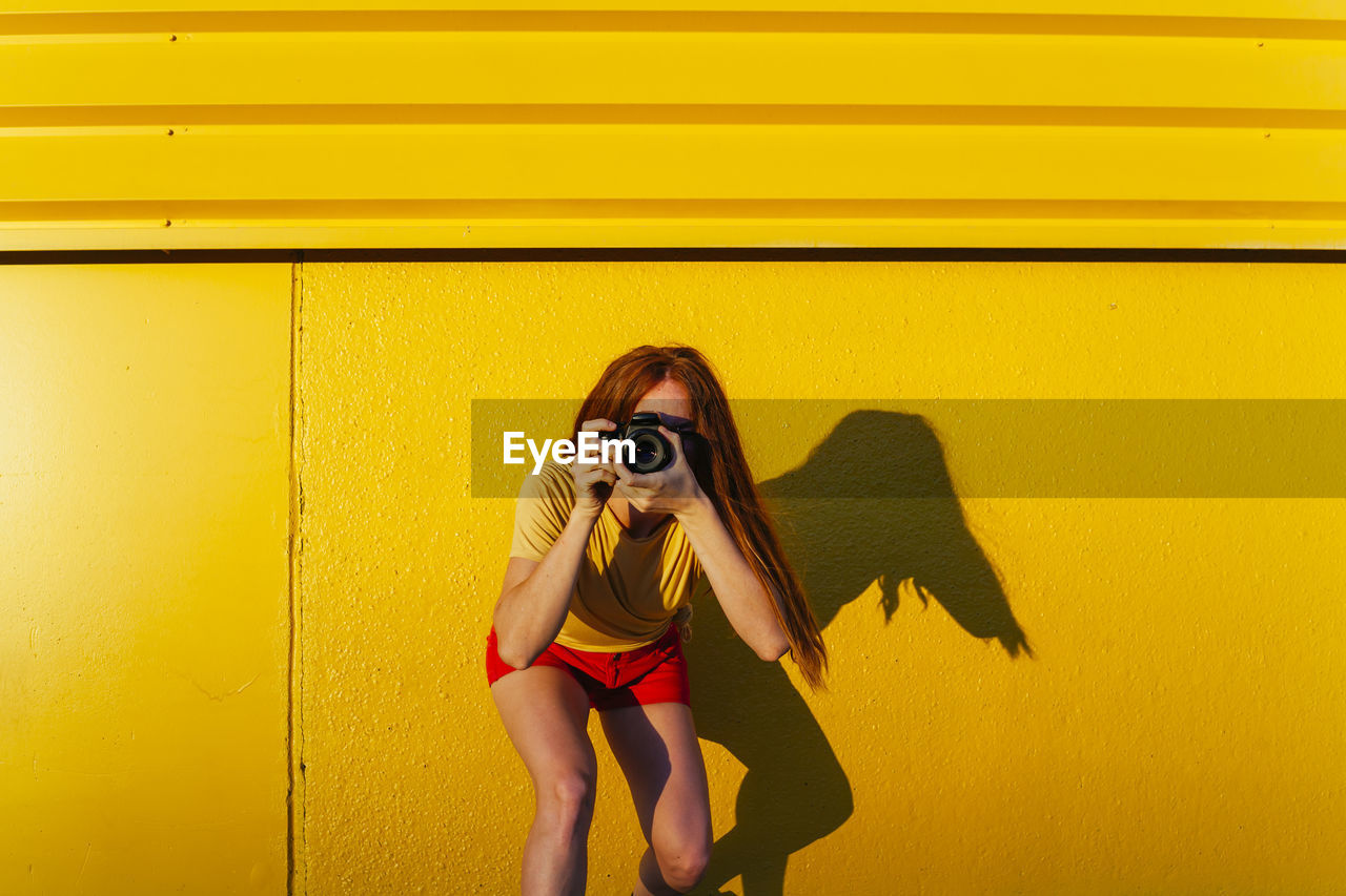 Woman photographing through camera while standing against yellow wall